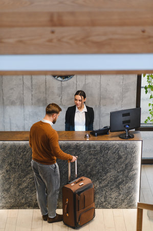 Traveler with luggage arriving at a modern hotel reception desk while receptionist provides customer serviceの写真素材