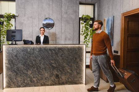 Business traveler pulling luggage, arriving at a stylish hotel lobby with a friendly female receptionist standing behind the counterの写真素材
