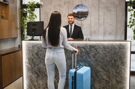 Guest standing with luggage at a modern hotel reception desk, interacting with a friendly, uniformed male conciergeの写真素材
