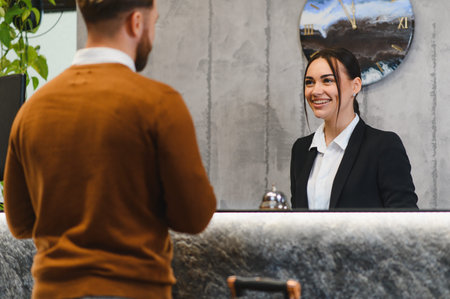 Friendly hotel receptionist smiling and helping a male guest at the reception desk, providing hospitality and customer serviceの写真素材