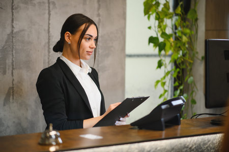 Professional woman working at a modern hotel reception, holding a clipboard and providing customer service with a welcoming expressionの写真素材