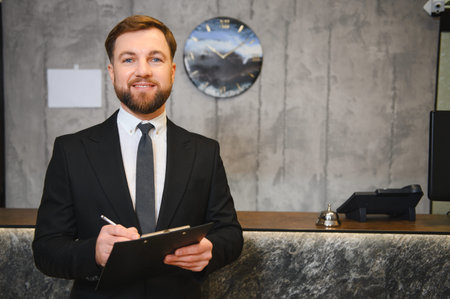Friendly male receptionist wearing a suit and writing on a clipboard, providing professional hospitality services in a modern lobbyの写真素材