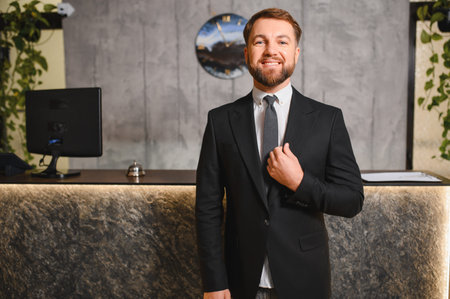 Smiling man working as a hotel receptionist or concierge, standing professionally at a modern reception desk in a lobbyの写真素材