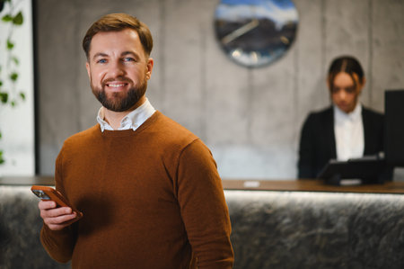 Happy man looking at camera, holding mobile phone in a modern hotel or business center with a blurred receptionist in backgroundの写真素材