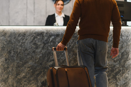 Man arriving at hotel, pulling a suitcase while approaching the reception desk, ready for check in and enjoying hospitalityの写真素材