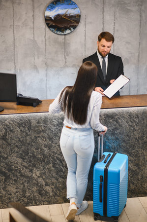 Female traveler with luggage completing check in process at a modern hotel front desk with a male receptionistの写真素材