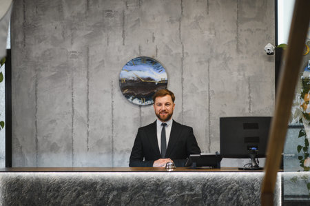 Friendly male receptionist wearing a suit and tie standing behind a modern hotel reception desk, ready to assist visitorsの写真素材