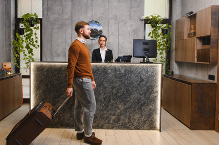 Man with luggage walking towards female receptionist at the modern hotel lobby desk, engaging in check in processの写真素材