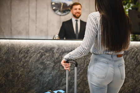Woman with luggage standing at a modern hotel reception desk, talking to a smiling male receptionist, hospitality conceptの写真素材
