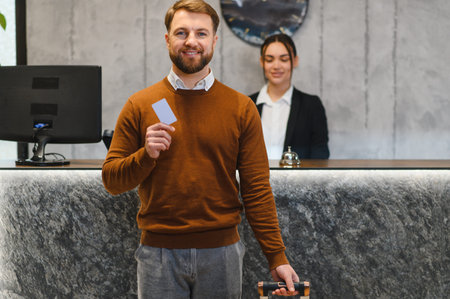 Happy traveler holding hotel key card and luggage at reception desk, smiling and ready for a comfortable, confident check in experienceの写真素材