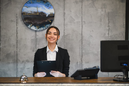 Smiling woman standing at a hotel reception desk, holding a clipboard and looking confident. Providing professional hospitality serviceの写真素材