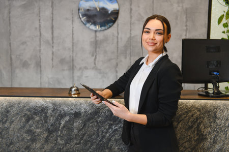 Smiling hotel receptionist standing at modern hotel front desk, holding a clipboard and looking at the cameraの写真素材