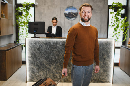 Smiling man pulling luggage at contemporary hotel lobby. Receptionist working at front desk, providing hospitality serviceの写真素材