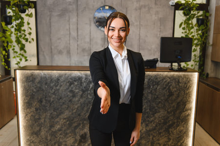Young woman standing at reception desk, extending hand, welcoming visitors with a friendly smile, showing hospitality conceptの写真素材