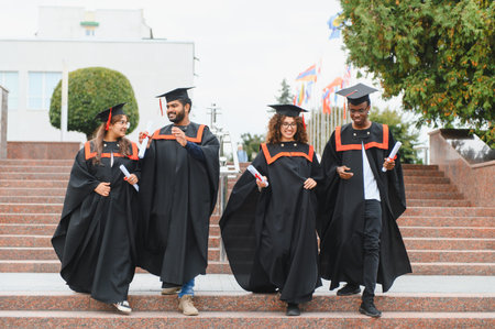 Graduates from various backgrounds walking down stairs on campus, holding diplomas and celebrating their academic achievementsの写真素材
