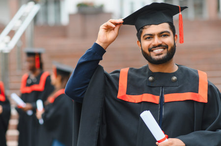 Happy Indian student celebrating graduation, wearing cap and gown, holding scroll outdoors with peers in backgroundの写真素材