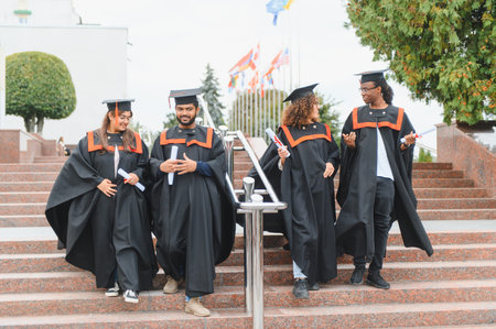 Diverse group of young graduates walking down stairs on campus, celebrating academic success and holding diplomasの写真素材