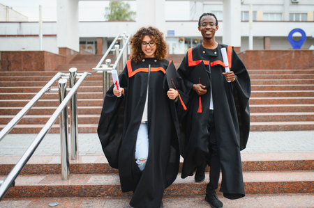 Happy recent graduates celebrating academic success, walking down university stairs with diplomas and cap and gown attireの写真素材