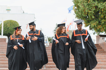 Group of diverse university students wearing academic gowns and caps, holding diplomas, and walking down stairs after graduation ceremonyの写真素材
