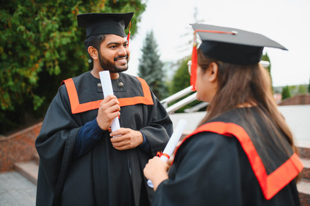 Two Indian university graduates happily discussing after receiving their diplomas, wearing academic cap and gownの写真素材
