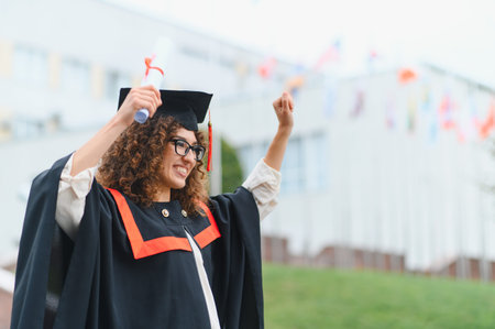 Graduating woman celebrating academic success, holding her diploma with joy and raising a fist in triumph outside a university buildingの写真素材
