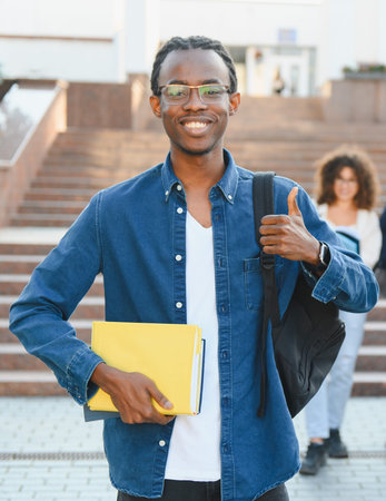 African american student smiling, holding books, giving a thumb up sign in front of a university buildingの写真素材