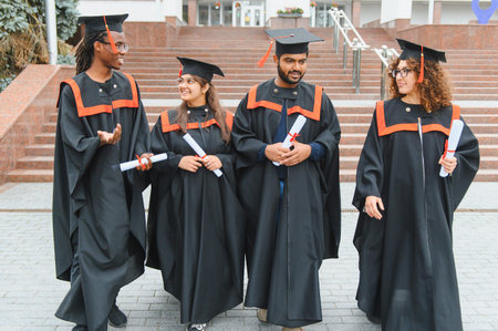 Group of happy multicultural students wearing academic gowns and caps, holding diplomas, celebrating graduation day on campusの写真素材