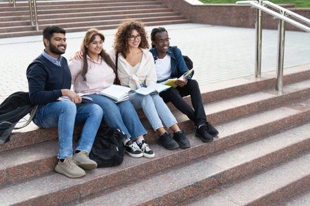 Group of diverse university students sitting on stairs, reading books, and smiling while discussing studies outdoorsの写真素材