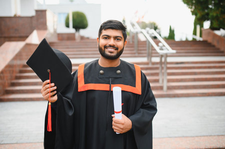Indian male graduate student smiling, holding a diploma and graduation cap, standing on a university campus staircaseの写真素材