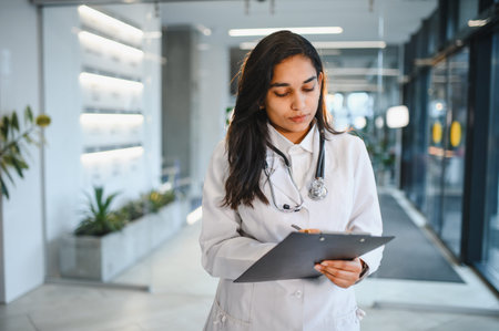 Medical professional standing in a modern clinic corridor, holding a clipboard and pen while reviewing healthcare notesの写真素材
