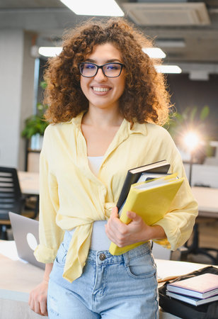 Young woman with curly hair and glasses holding textbooks, standing in a brightly lit learning environmentの写真素材