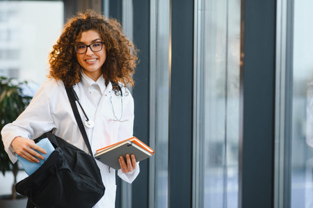 Young female medical student wearing a lab coat and stethoscope, carrying books and a bag, smiling at the cameraの写真素材
