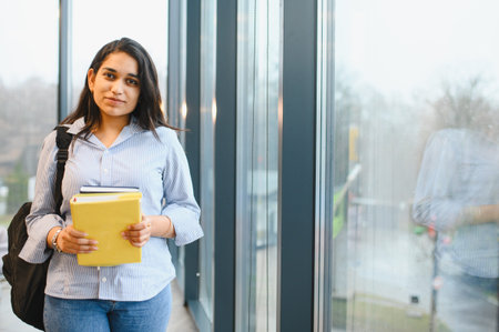 Indian university student standing with books and backpack in a modern campus building, representing education and learningの写真素材