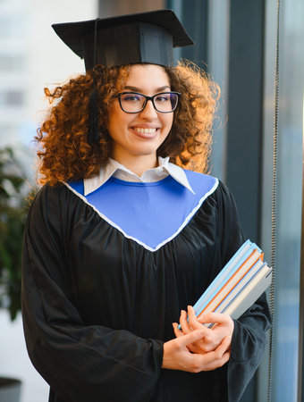 Female graduate student in academic dress and cap, holding books and smiling, celebrating her academic successの写真素材
