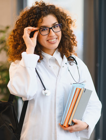 Young female medical student smiling, adjusting eyeglasses, holding textbooks, and wearing a lab coat with a stethoscopeの写真素材