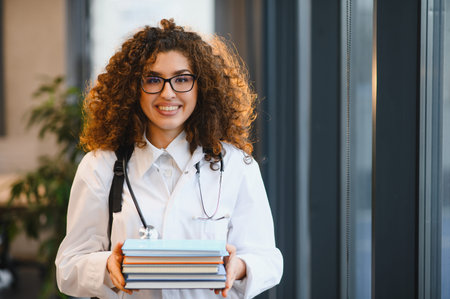 Young female medical student wearing lab coat and stethoscope, holding books and smiling, ready for her healthcare studiesの写真素材