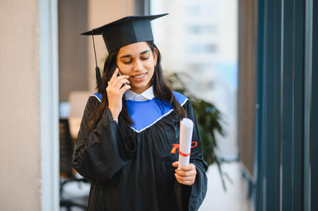 Indian graduate student talking on phone, holding university diploma, celebrating education achievement and bright futureの写真素材