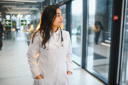 Indian doctor wearing a white lab coat and stethoscope, walking confidently in a bright clinic corridor, representing healthcare and professionalismの写真素材
