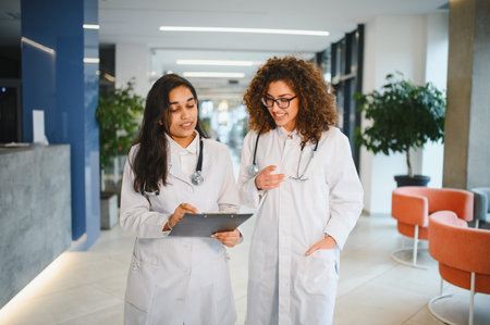 Two multiracial women doctors in white coats reviewing information on a clipboard, collaborating in a modern healthcare facilityの写真素材