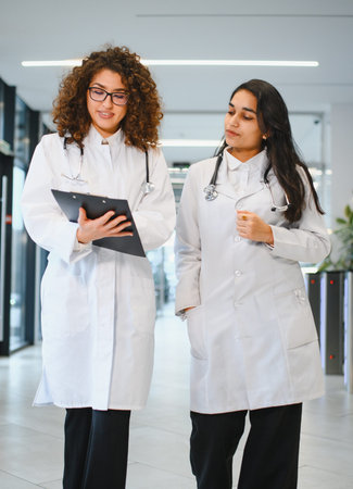 Two doctors wearing lab coats and stethoscopes walking and discussing patient records on a clipboard in a modern clinicの写真素材
