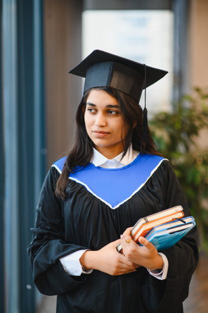 Indian woman student wearing a graduation cap and gown, holding books, representing successful academic achievement and future careerの写真素材