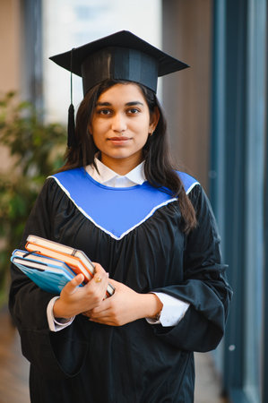 Young Indian woman wearing graduation cap and gown, holding educational books. Representing success and academic achievementの写真素材