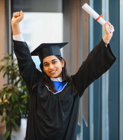 Indian female student graduation wearing cap and gown, smiling brightly while holding her diploma, celebrating academic successの写真素材