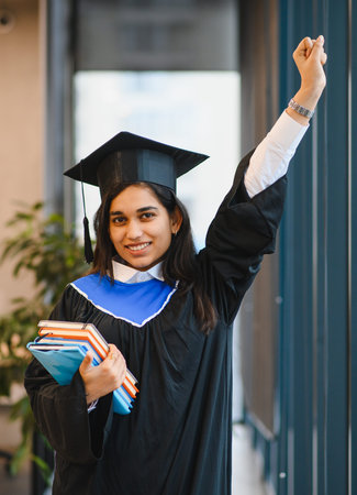 Happy Indian student in graduation gown and cap raising an arm in celebration, achieving academic goals and educationの写真素材