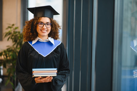 Smiling female graduate student in cap and gown holding books, celebrating academic achievement and a bright futureの写真素材
