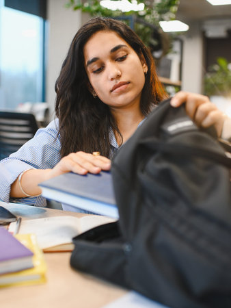 Young Indian student preparing for classes, packing books in a backpack at a university library or study areaの写真素材