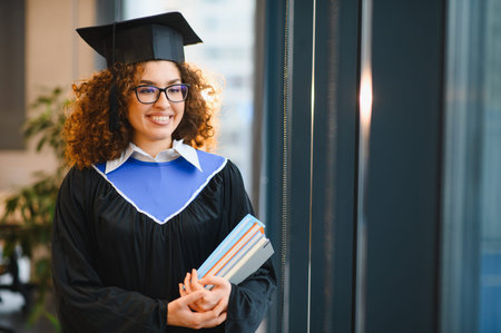 Female student smiling, wearing a graduation cap and gown, holding books, celebrating academic achievement and future successの写真素材