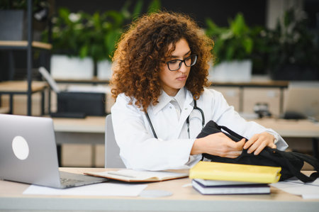Medical student woman wearing a lab coat and stethoscope, packing her bag after studying with a laptop and booksの写真素材