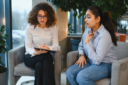 Female psychologist writing notes while listening to a young Indian woman patient discussing her mental health during a therapy sessionの写真素材