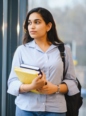 Young Indian woman carrying books and a backpack, standing indoors, looking away while focusing on her academic futureの写真素材